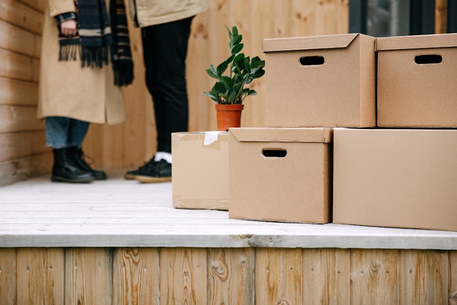 Cardboard boxes stacked on a wooden porch, symbolizing moving day with a potted plant nearby.